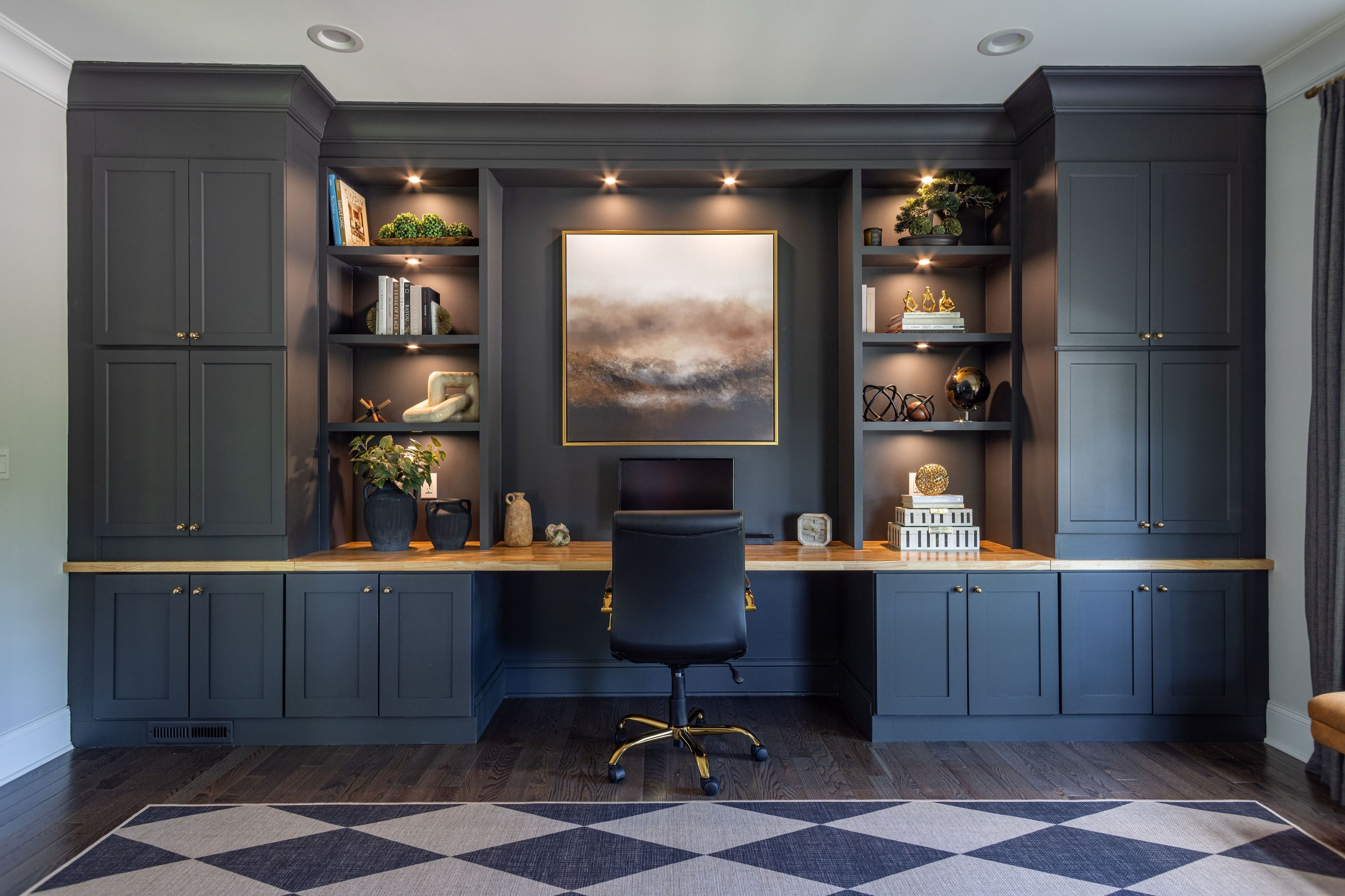 Home office featuring custom charcoal cabinetry and illuminated open shelving surrounding a wood desk and framed landscape artwork.