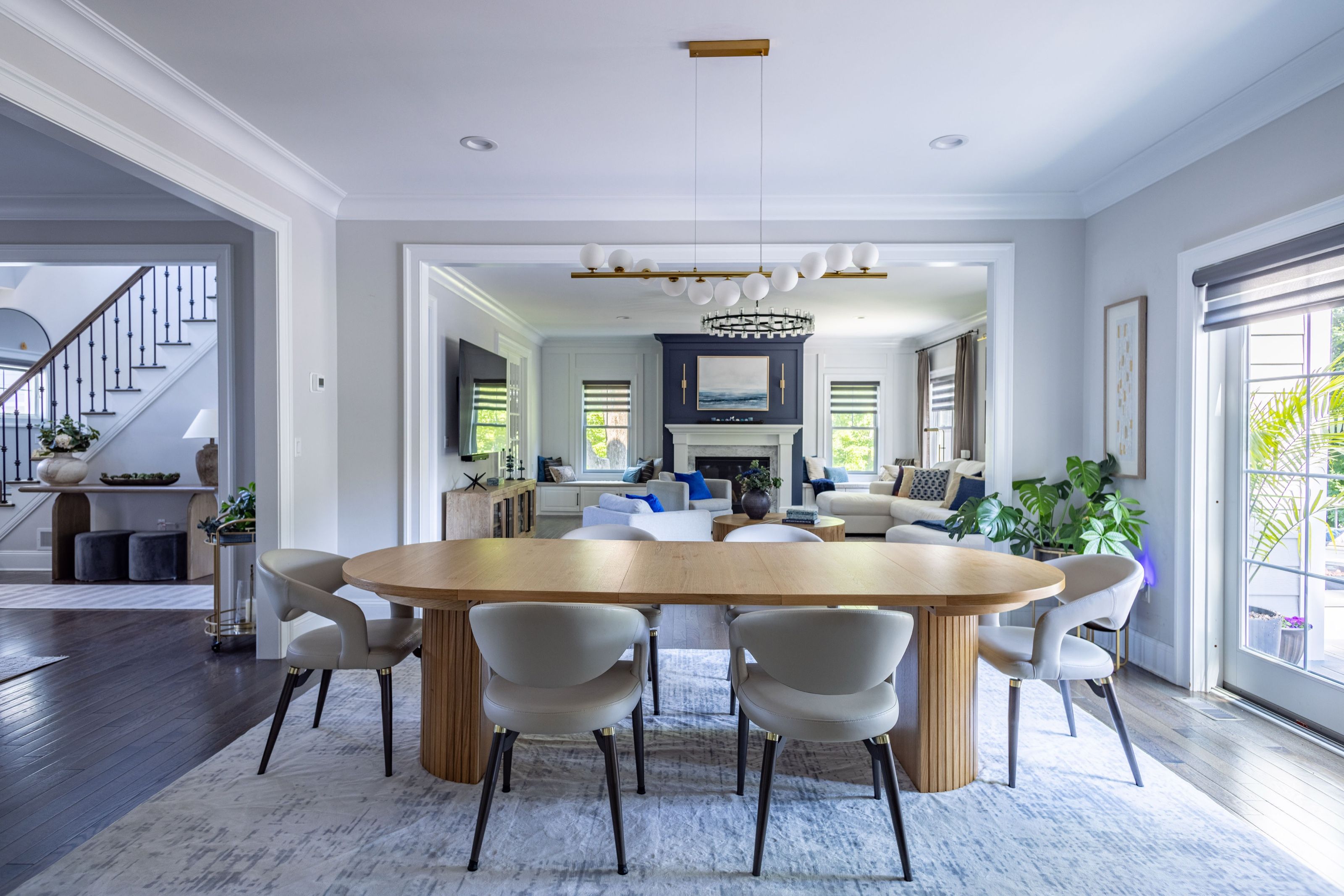 Dining room with an oval wood table with fluted legs, upholstered chairs, and a linear chandelier, opening to the family room.