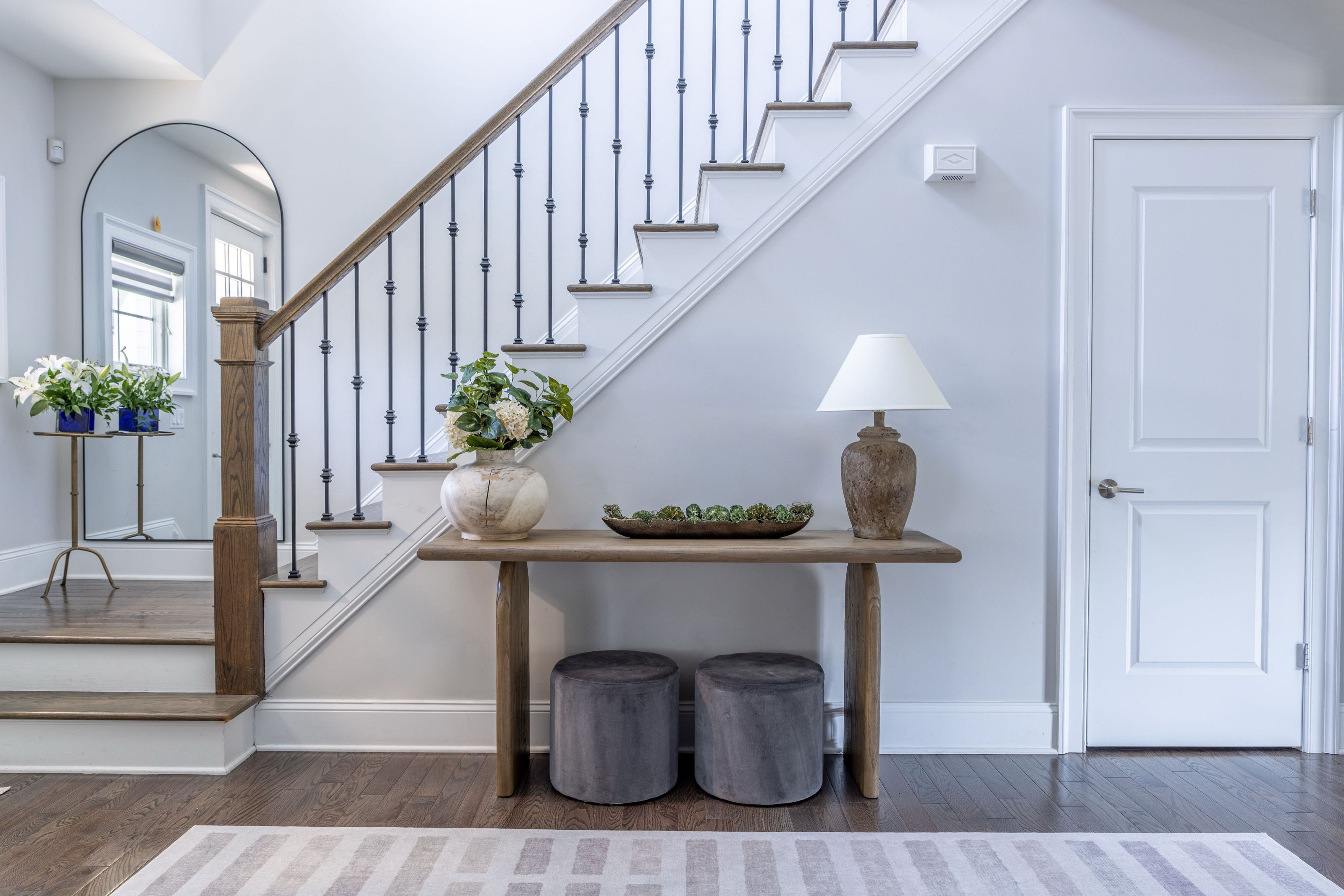 Entryway with a console table beneath the staircase, an arched mirror, and textured stools.