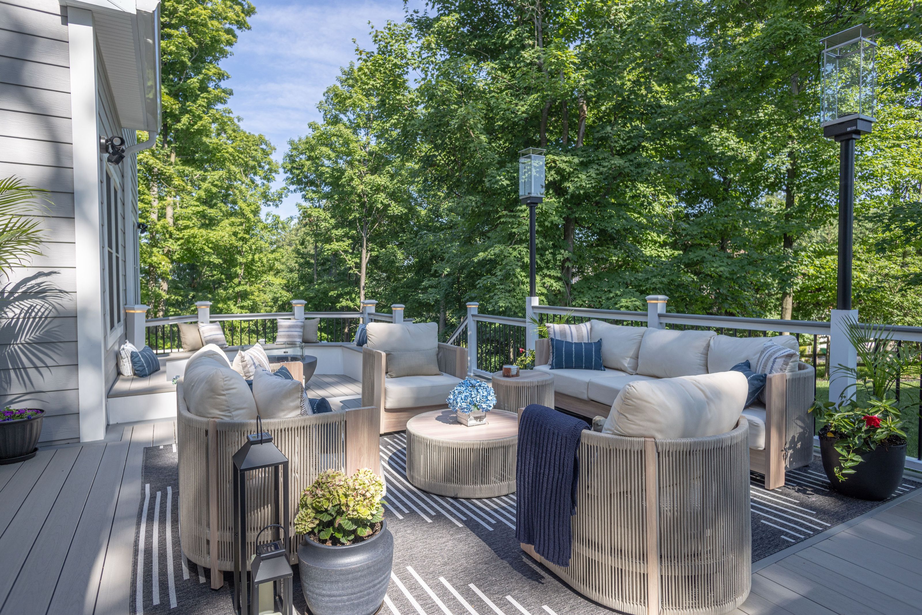 Outdoor living space with woven sofas and armchairs arranged around a low coffee table, surrounded by trees, and an outdoor firepit and seating area beyond.
