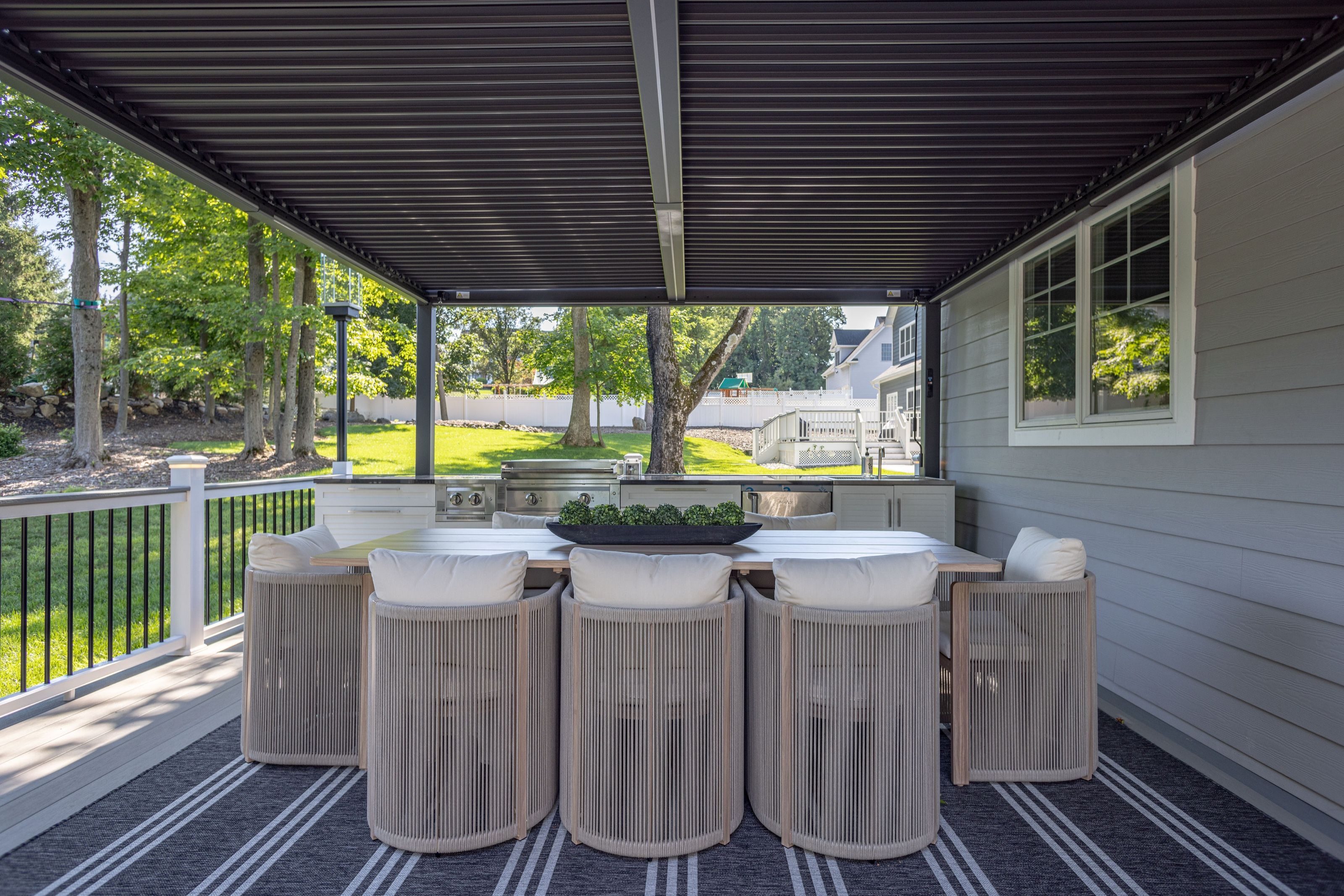 Outdoor dining area beneath a pergola, with a long wood table, woven chairs, an outdoor kitchen, and a woven rug.
