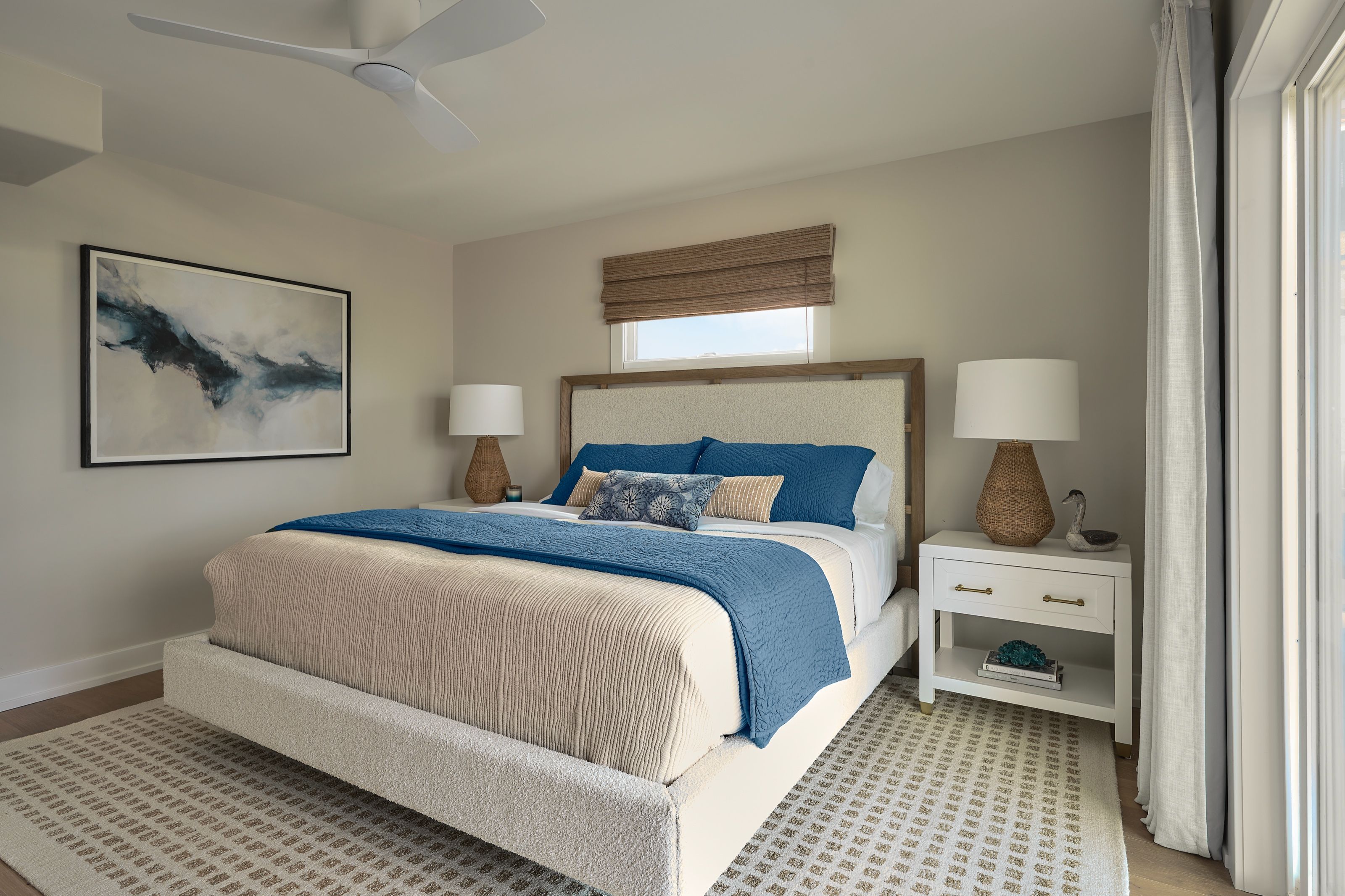 Primary bedroom with an upholstered bed, crisp white linens, navy throw, textured lumbar pillows, woven lamps, cream-and-tan wool rug, and abstract seascape artwork.