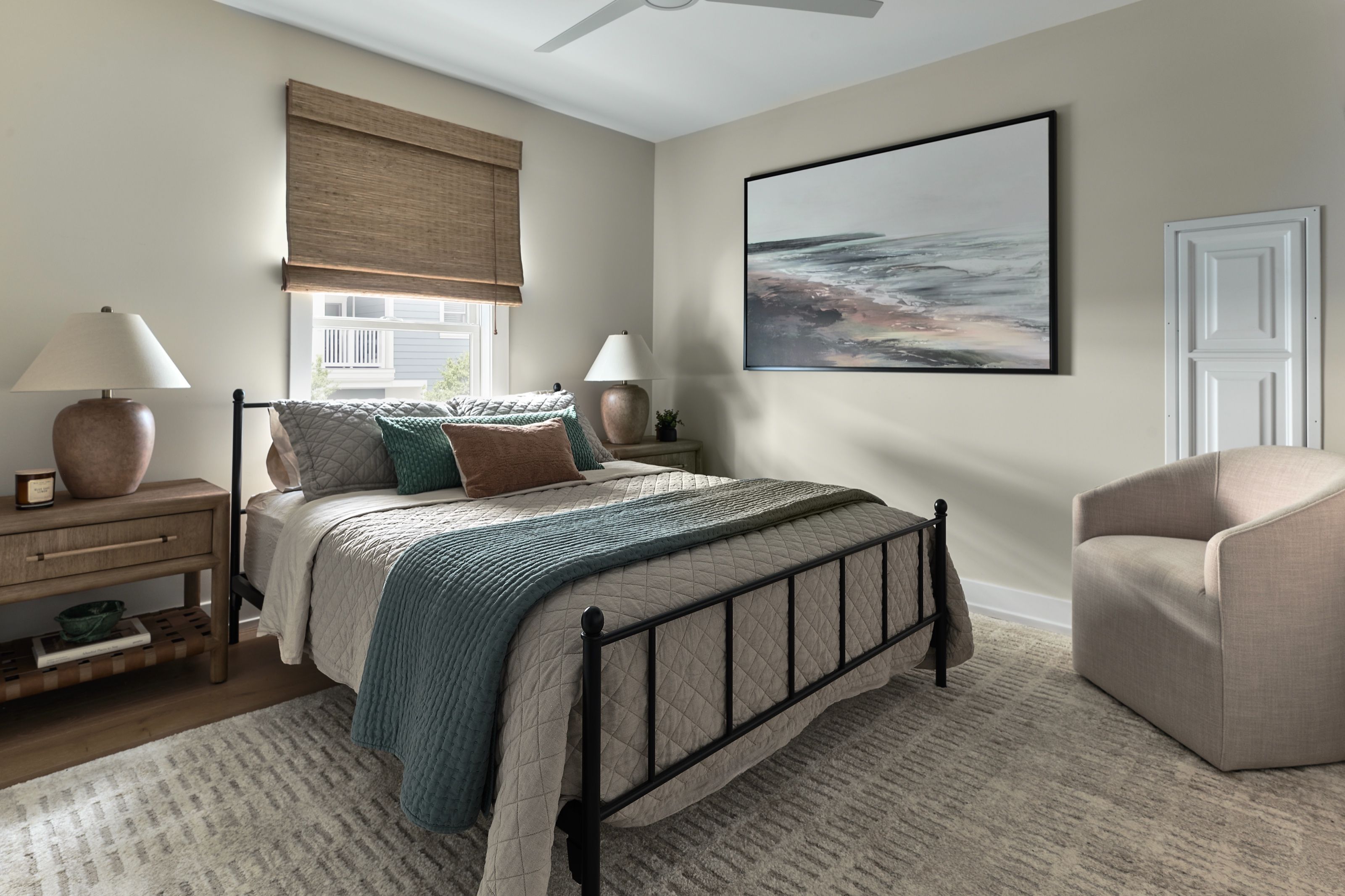 Guest bedroom with a black iron bed, tan quilt, teal coverlet, textured throw pillows, and abstract shoreline painting.