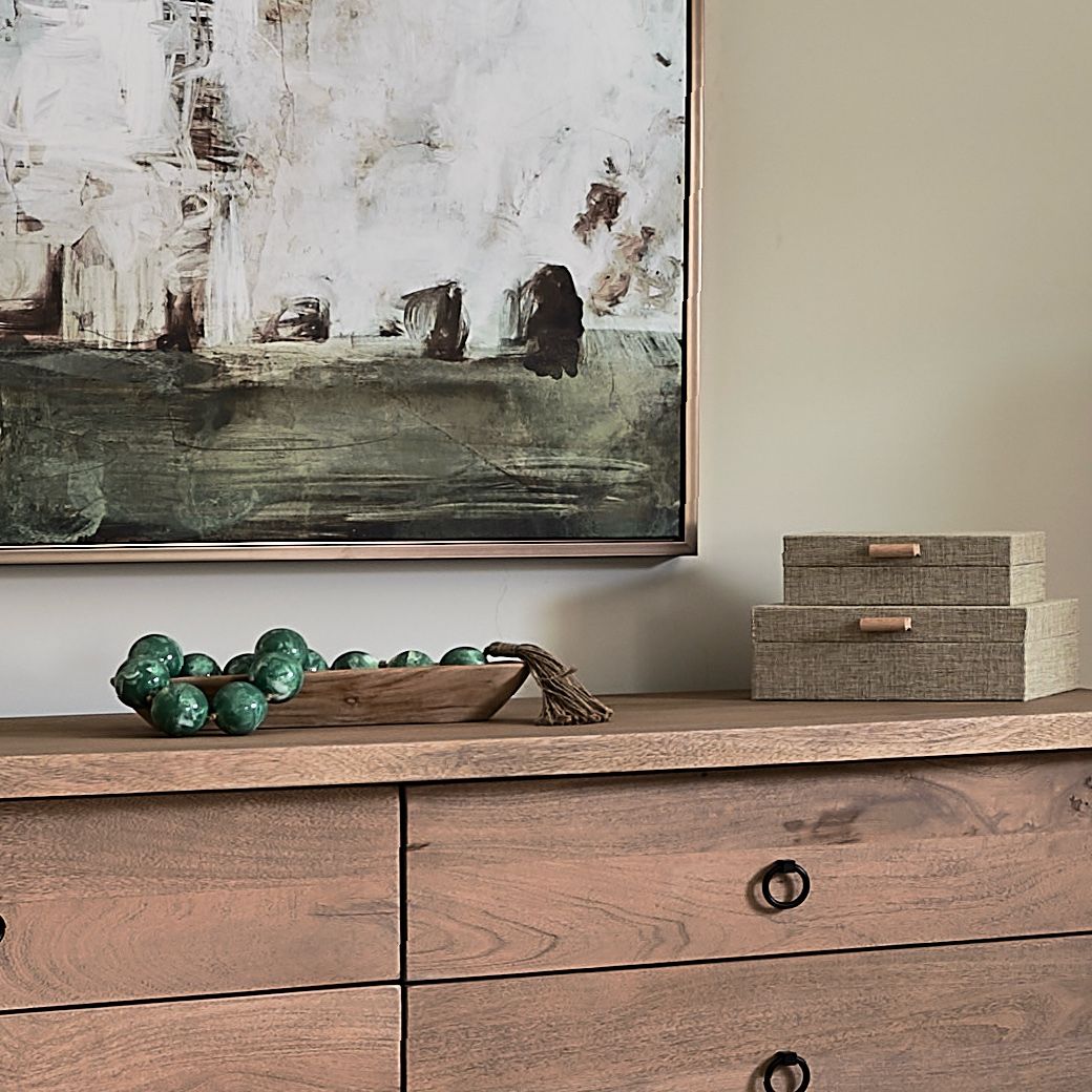 Bedroom dresser styled with a wooden bowl of green glass beads and woven boxes beneath an abstract painting.
