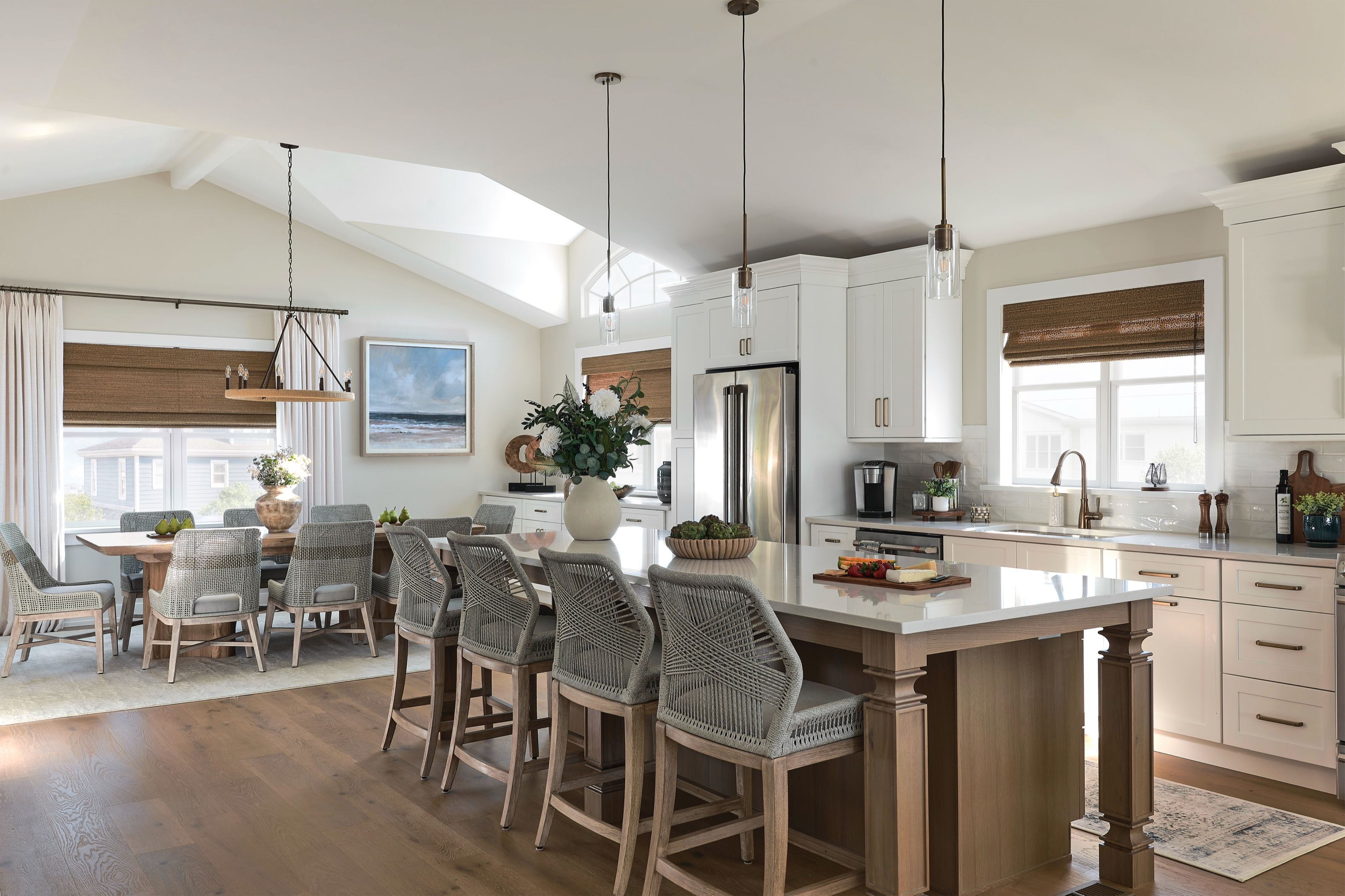 Open coastal kitchen with a wood island, woven counter stools, and custom woven wood window treatments, overlooking the dining area.