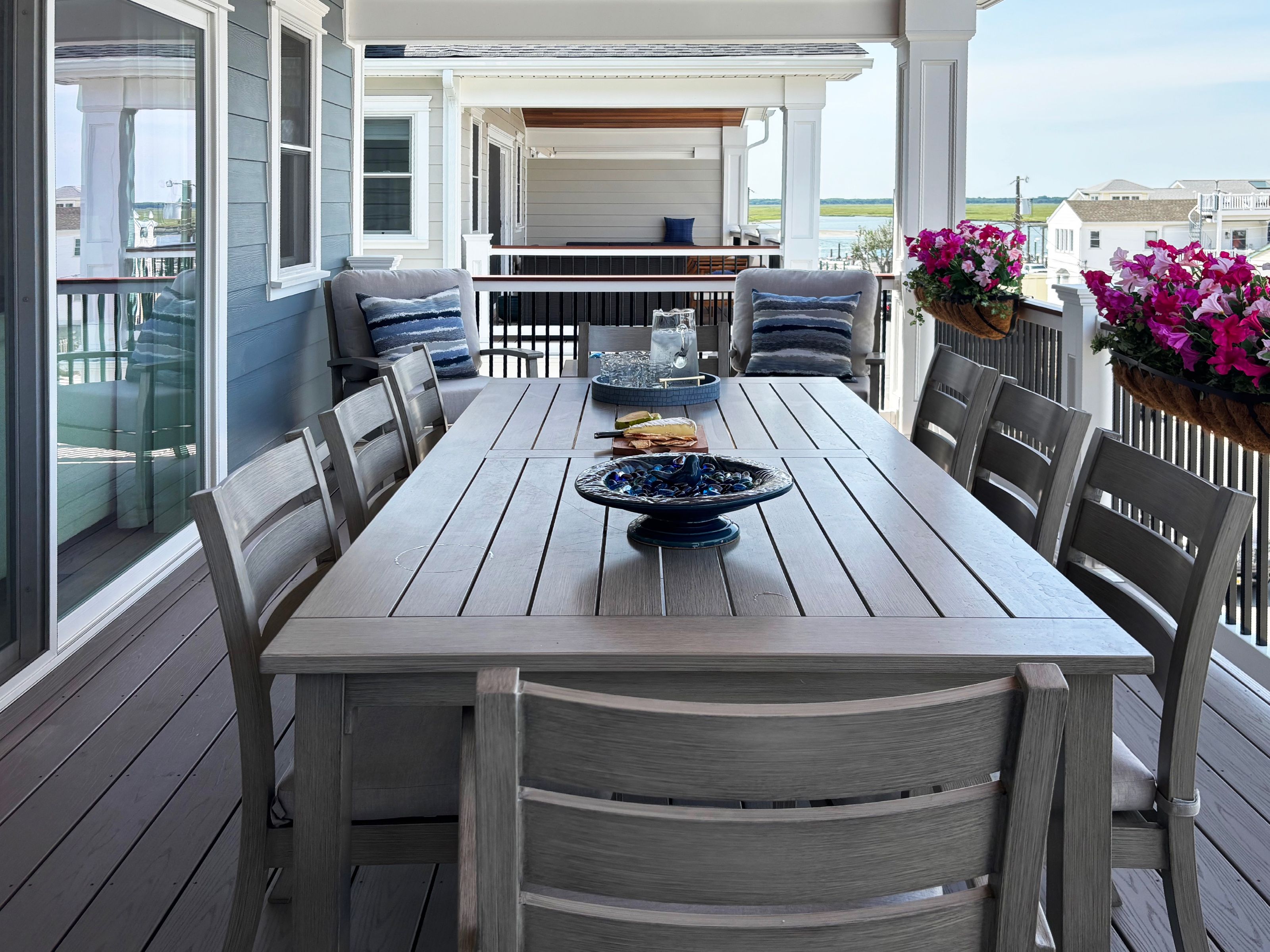 Covered coastal deck with a long gray dining table, flowering railing planters, and views toward the bay.