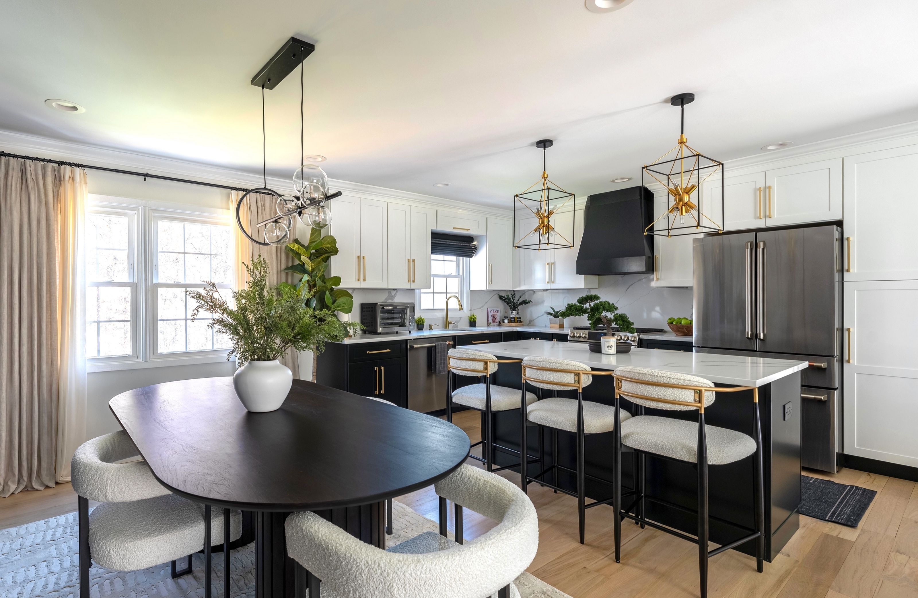 Renovated open kitchen and dining area with contrasting black and white cabinetry, an oval wood dining table, brass lantern pendants, and white oak floors in a Monmouth County, New Jersey home.